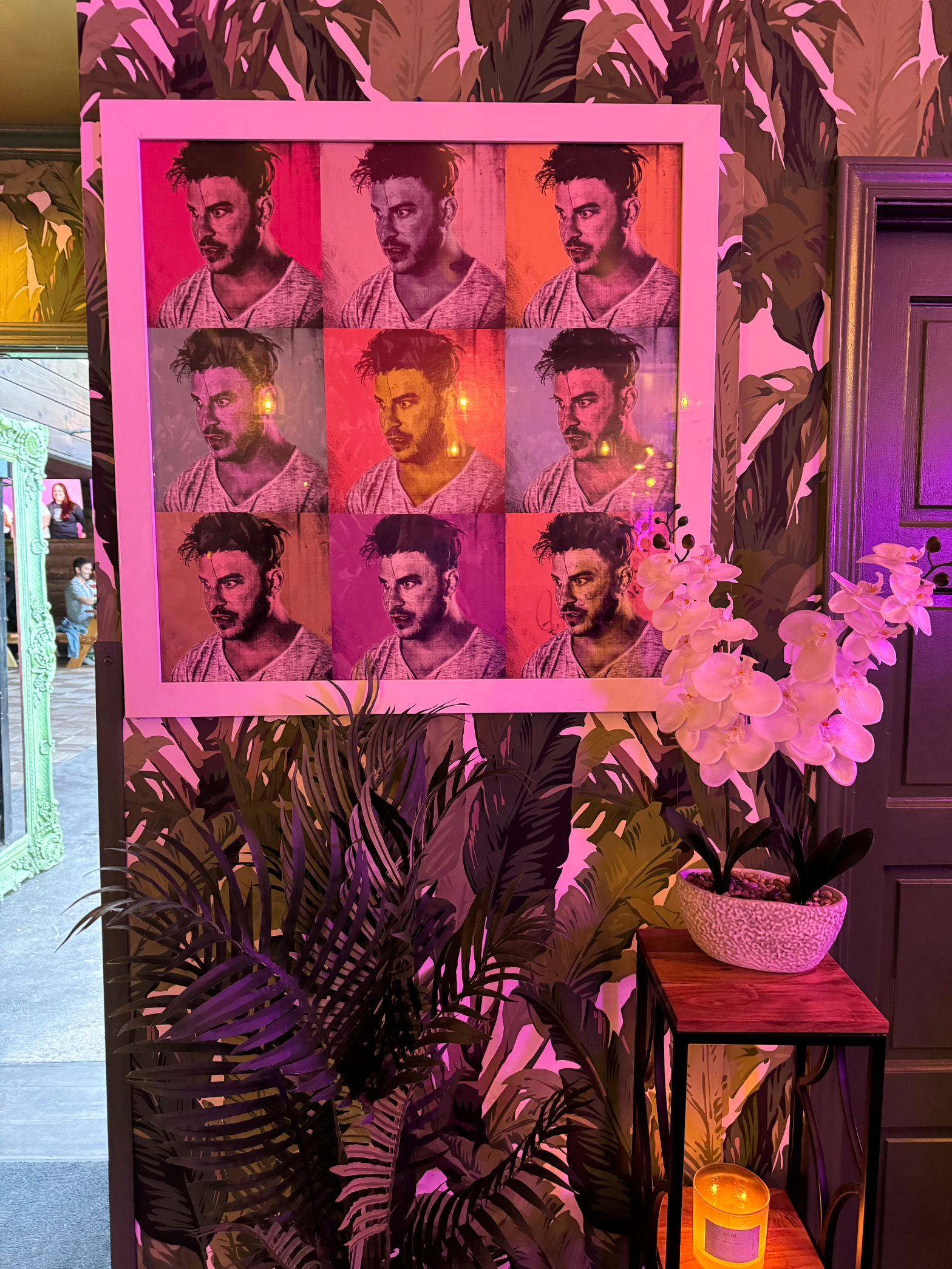 Artwork with repeated portraits above a plant and table with a candle and orchids, in a restaurant setting