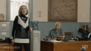 Two characters from "Schitt's Creek" in an office setting; one is standing and the other is sitting behind a desk