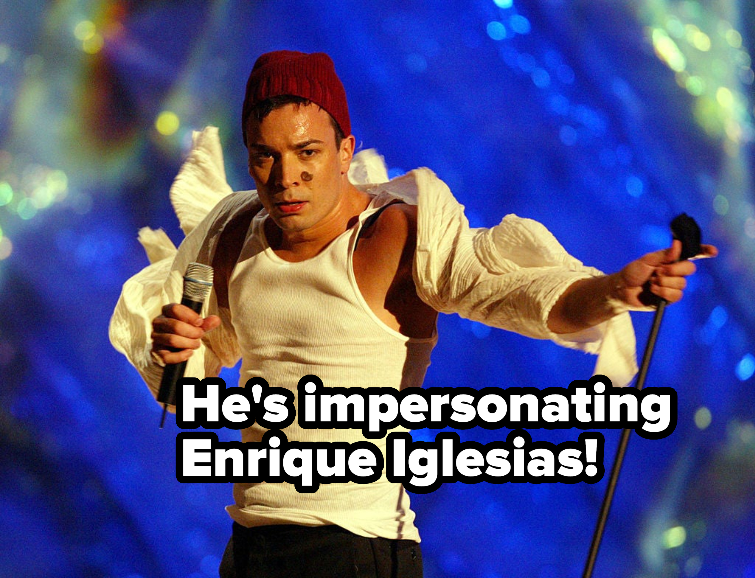 Performer on stage wearing a red beanie and casual white top with sleeves removed, singing into a microphone