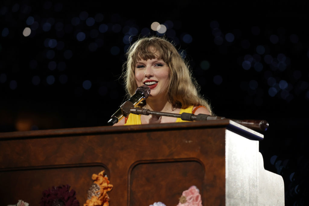 Taylor Swift is speaking into a microphone at a piano with floral decorations