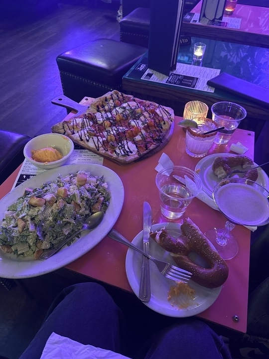 Assorted dishes on a table including a salad, a grilled platter, pretzel with dip, and beverages, viewed from a diner&#x27;s perspective