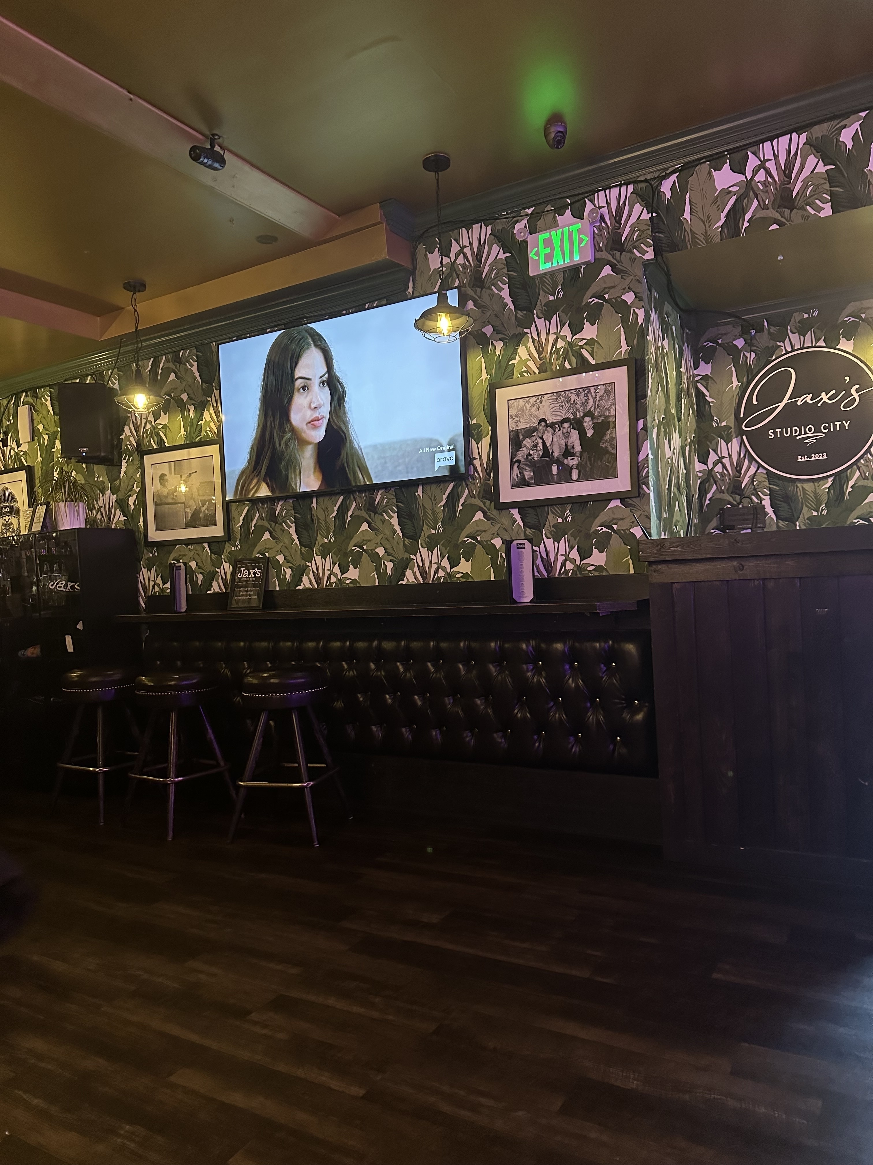 Interior of a bar with tropical wallpaper, framed pictures on the wall, and a TV screen displaying a woman