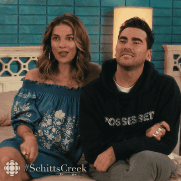 Two characters from Schitt's Creek sitting side by side, one in a floral top and one in a sweatshirt
