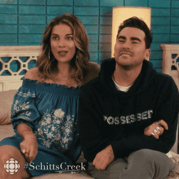 Two characters from Schitt&#x27;s Creek sitting side by side, one in a floral top and one in a sweatshirt