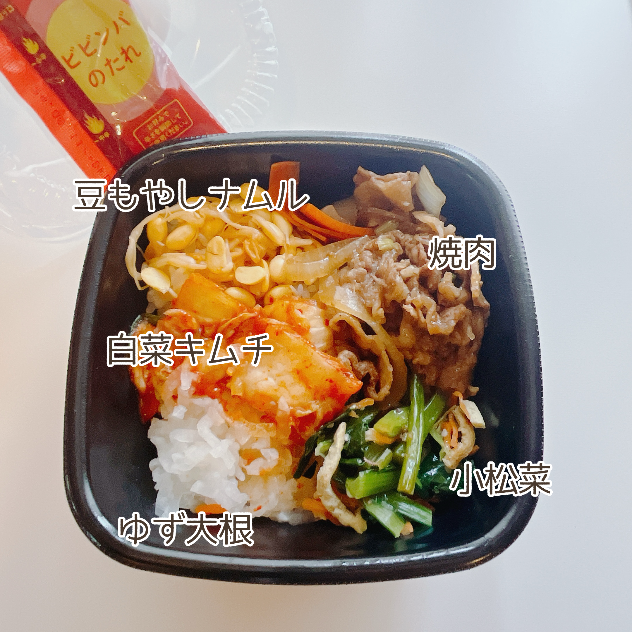 Top view of a bento box with rice, beef, noodles, and vegetables, accompanied by a drink. Text labels in Japanese