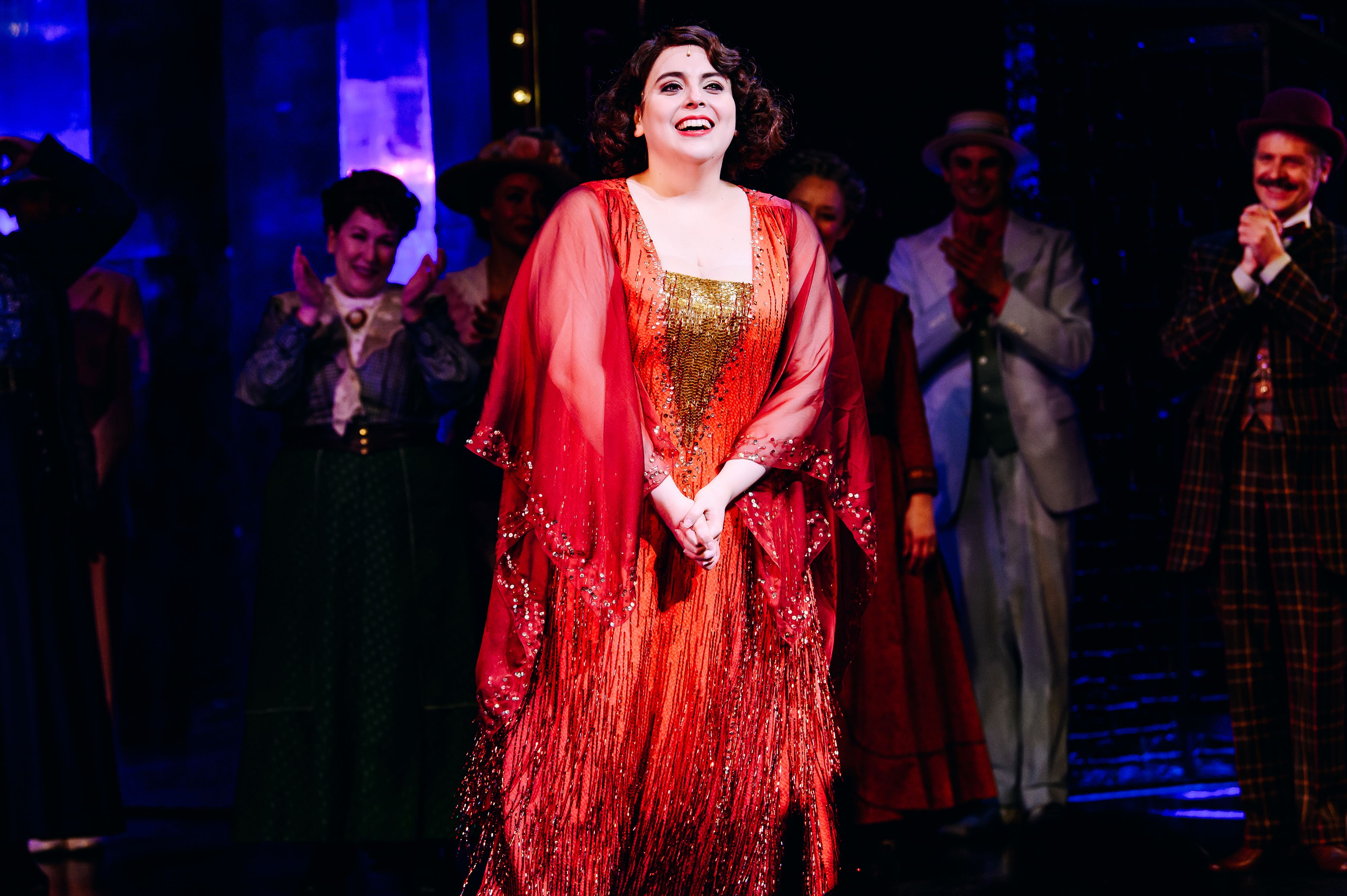 Woman in stage costume with sequins and a sheer red shawl, smiling on theater stage with cast in background