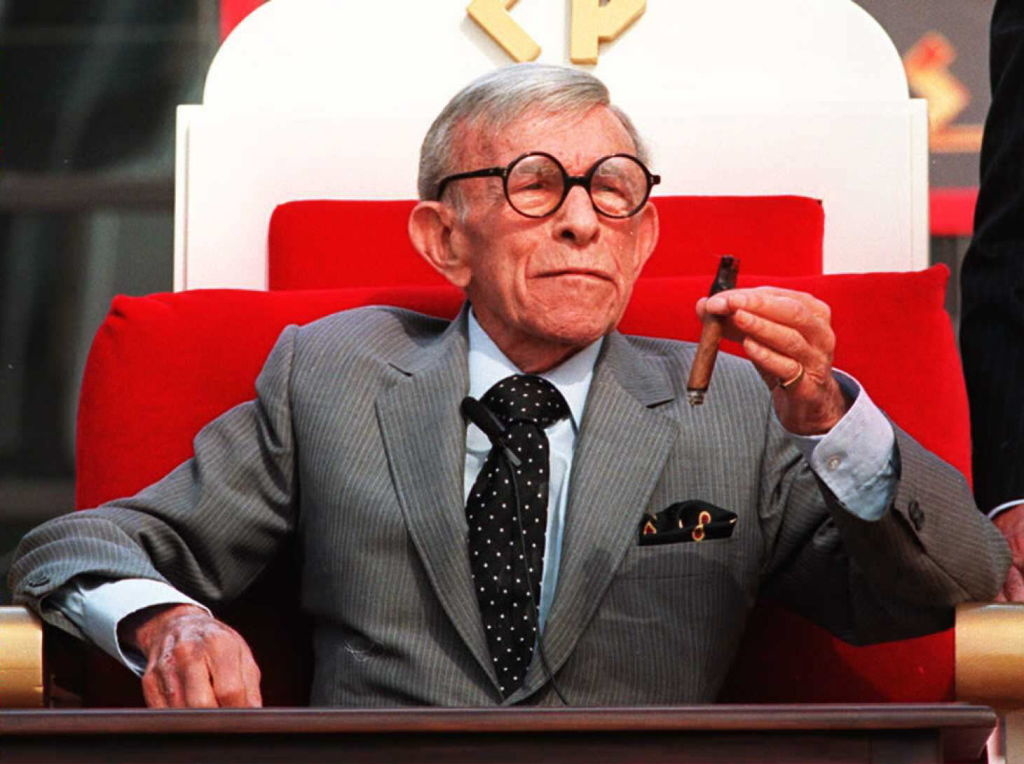 George Burns in a gray suit, seated with a cigar, at a celebratory event