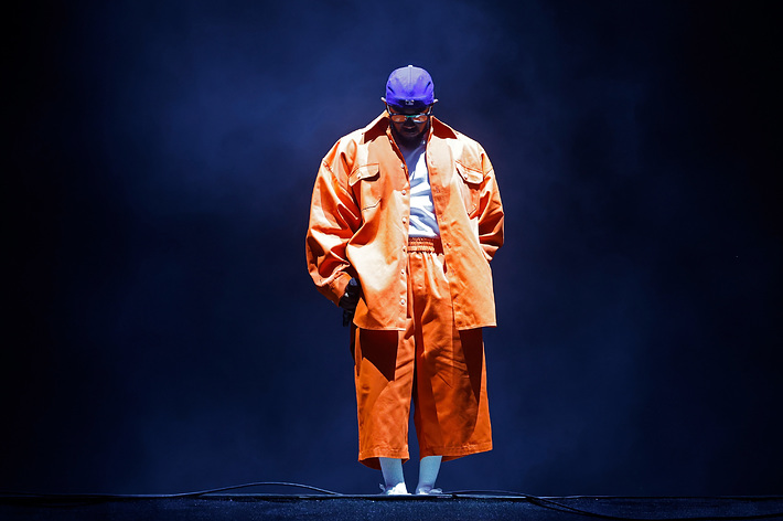 Musician in an oversized orange jacket and blue cap stands on stage under a spotlight