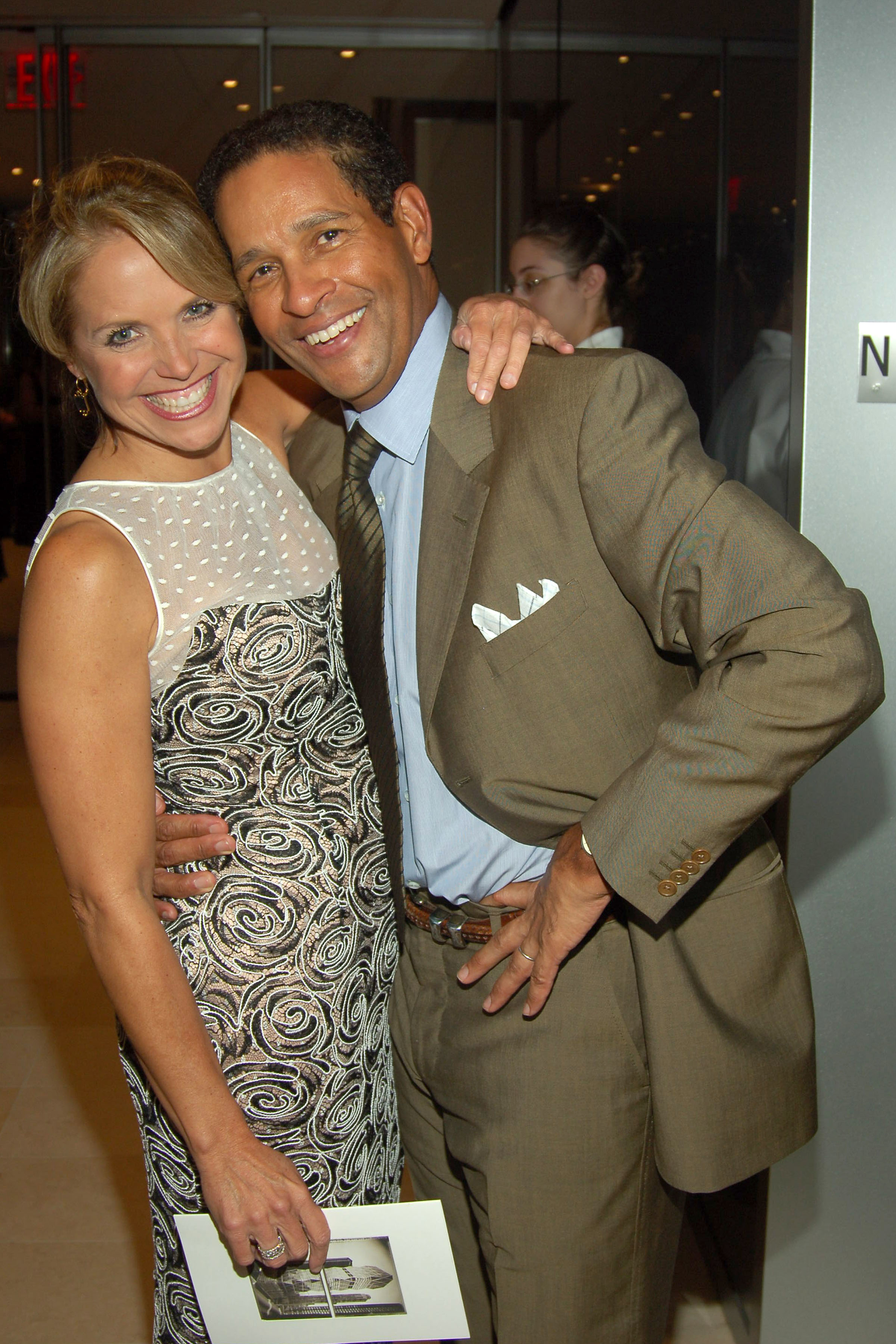 Katie Couric and Bryant Gumbel at an event, both smiling. Katie is in a patterned dress and Bryant is in a suit with a pocket square