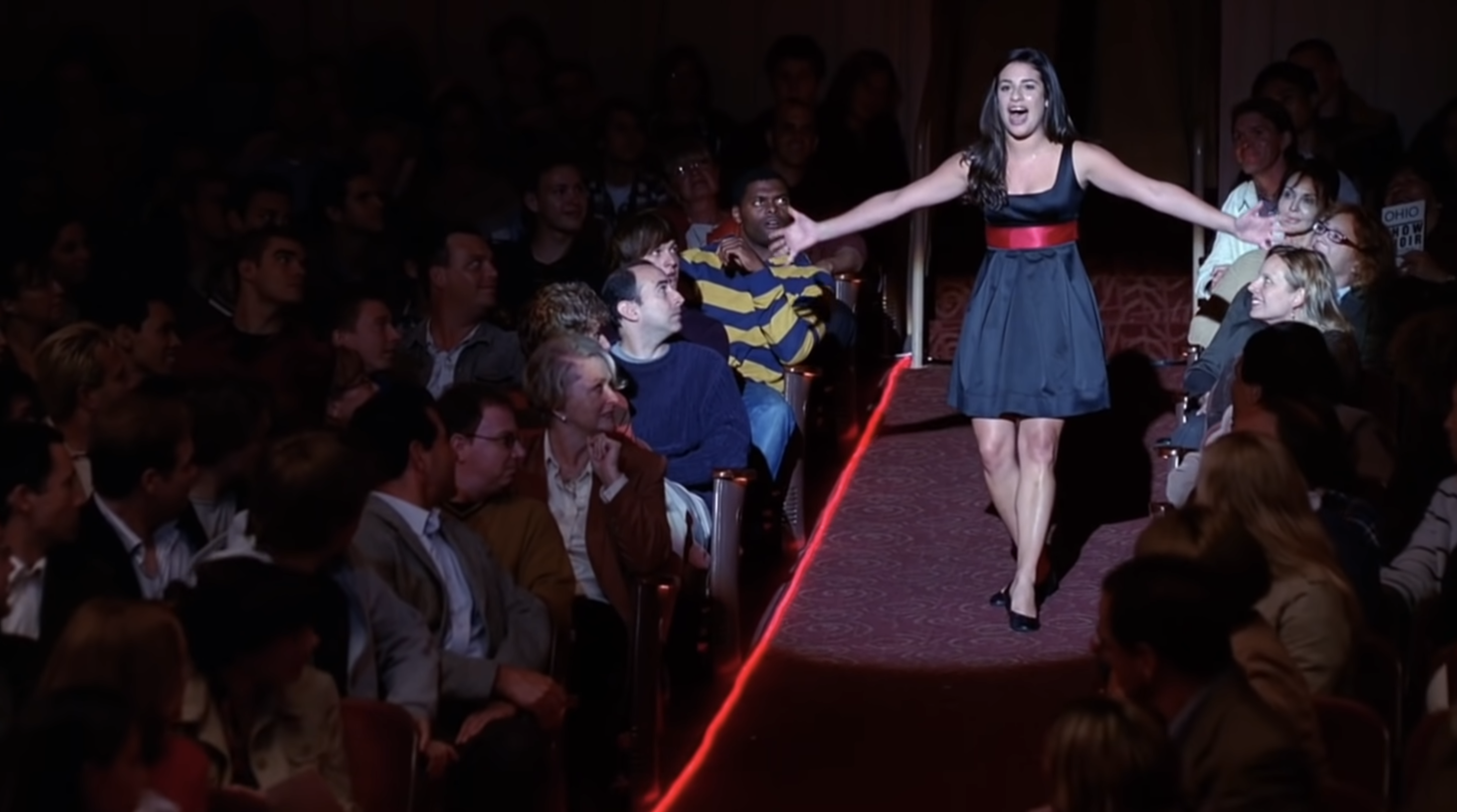Woman in a fashion show reaching out to audience wearing a chic dress with a red belt