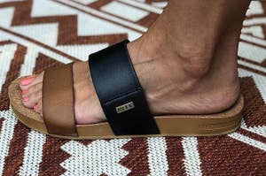Foot wearing a two-tone slide sandal with a "REEF" label on the strap, on a patterned floor