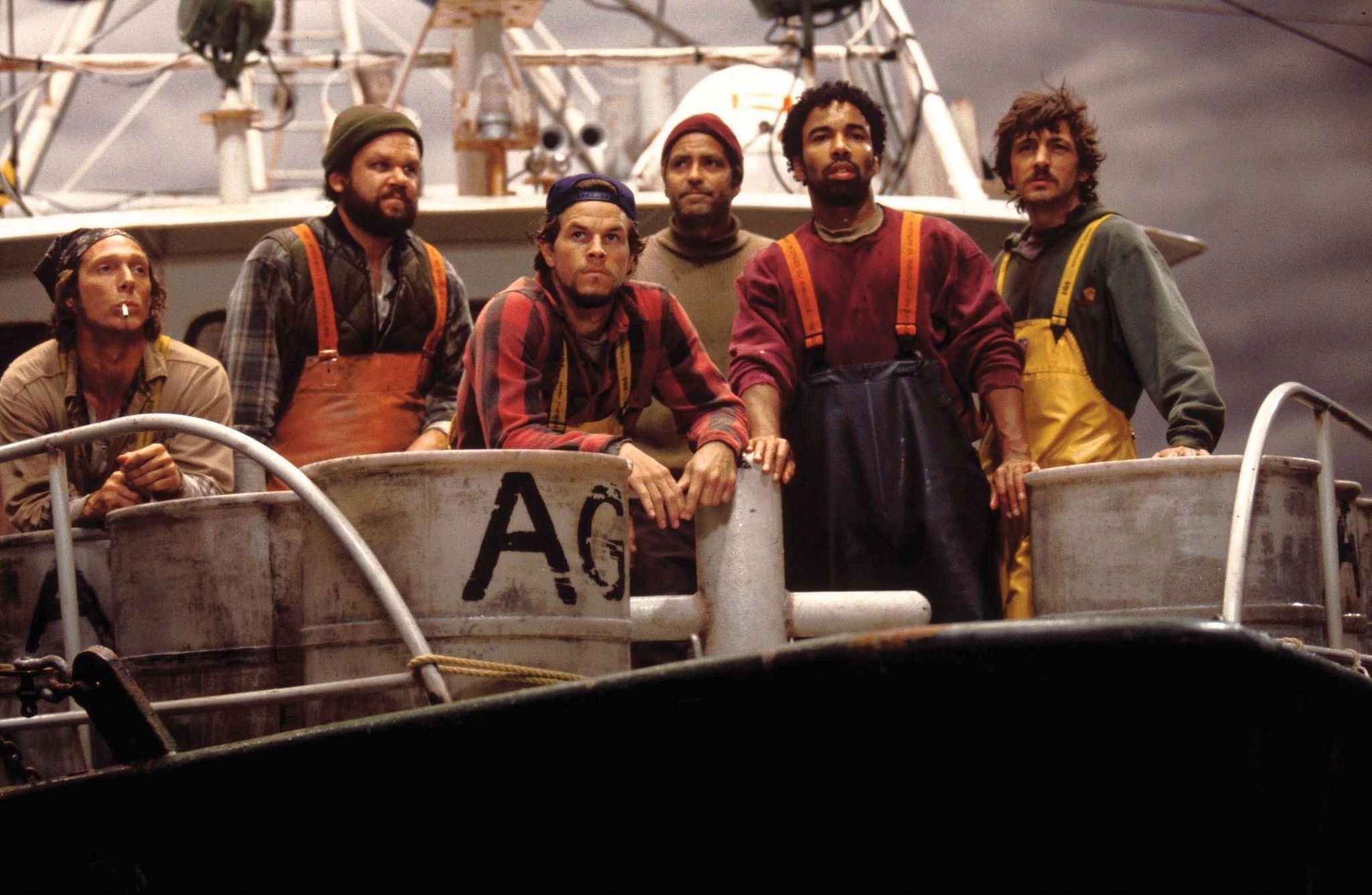 Six actors portraying fishermen in rugged gear on a boat, looking intently beyond the camera