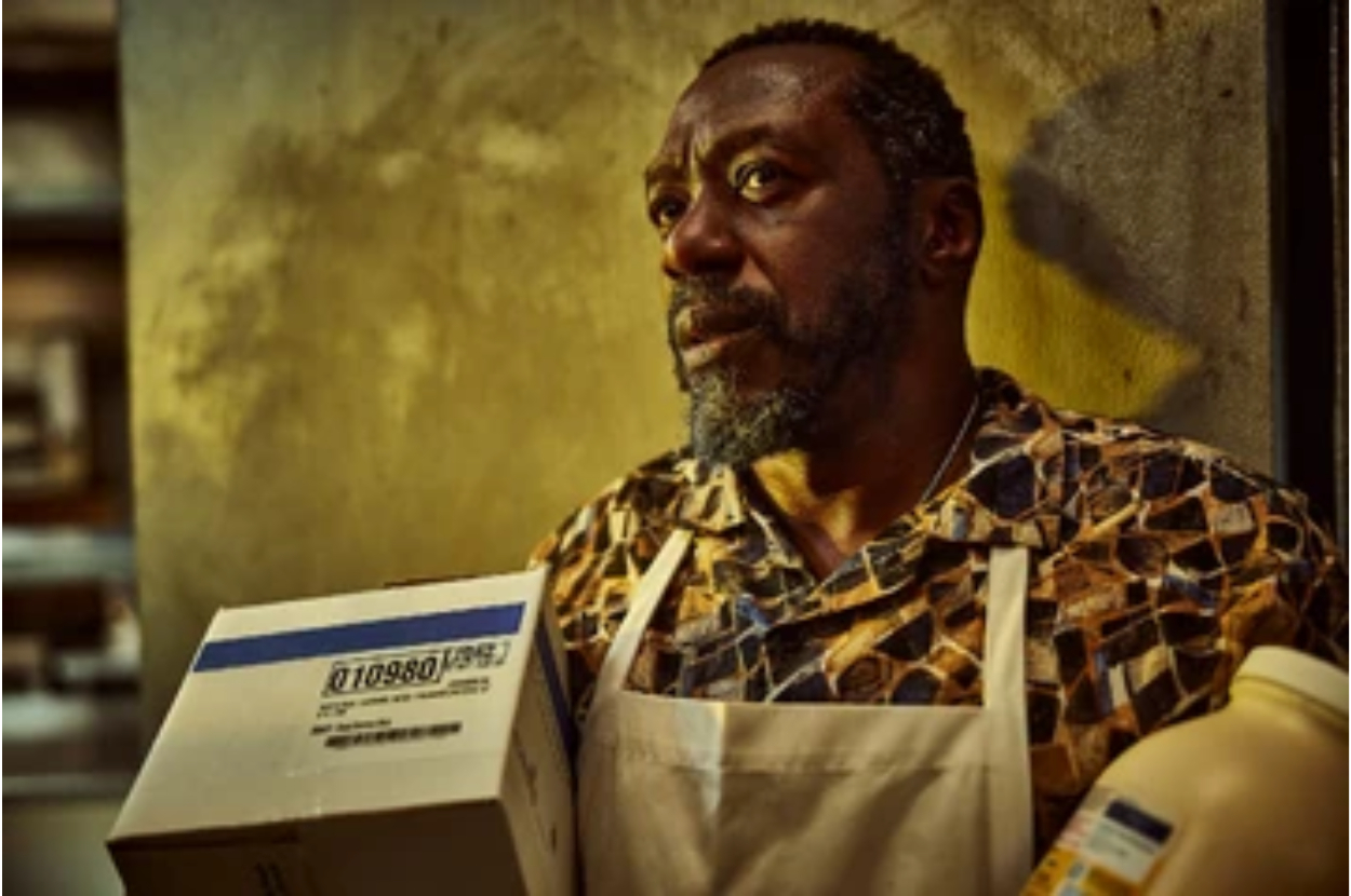 Man in a patterned shirt and apron holds a box, looking pensive; setting appears to be a storeroom