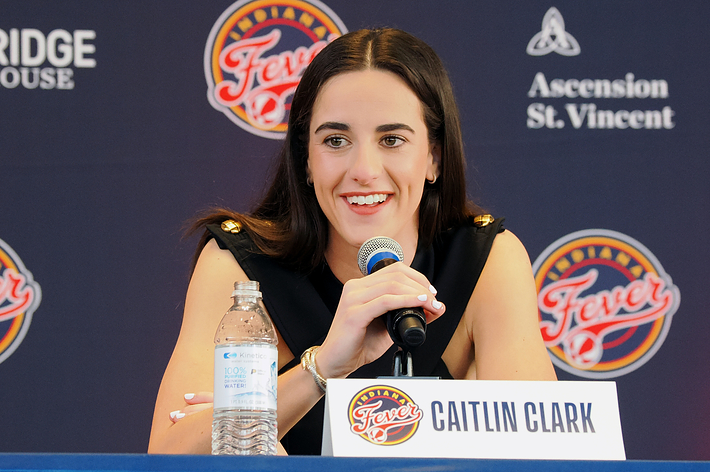 Caitlin Clark speaking into a microphone at a press conference with basketball team logos in the background
