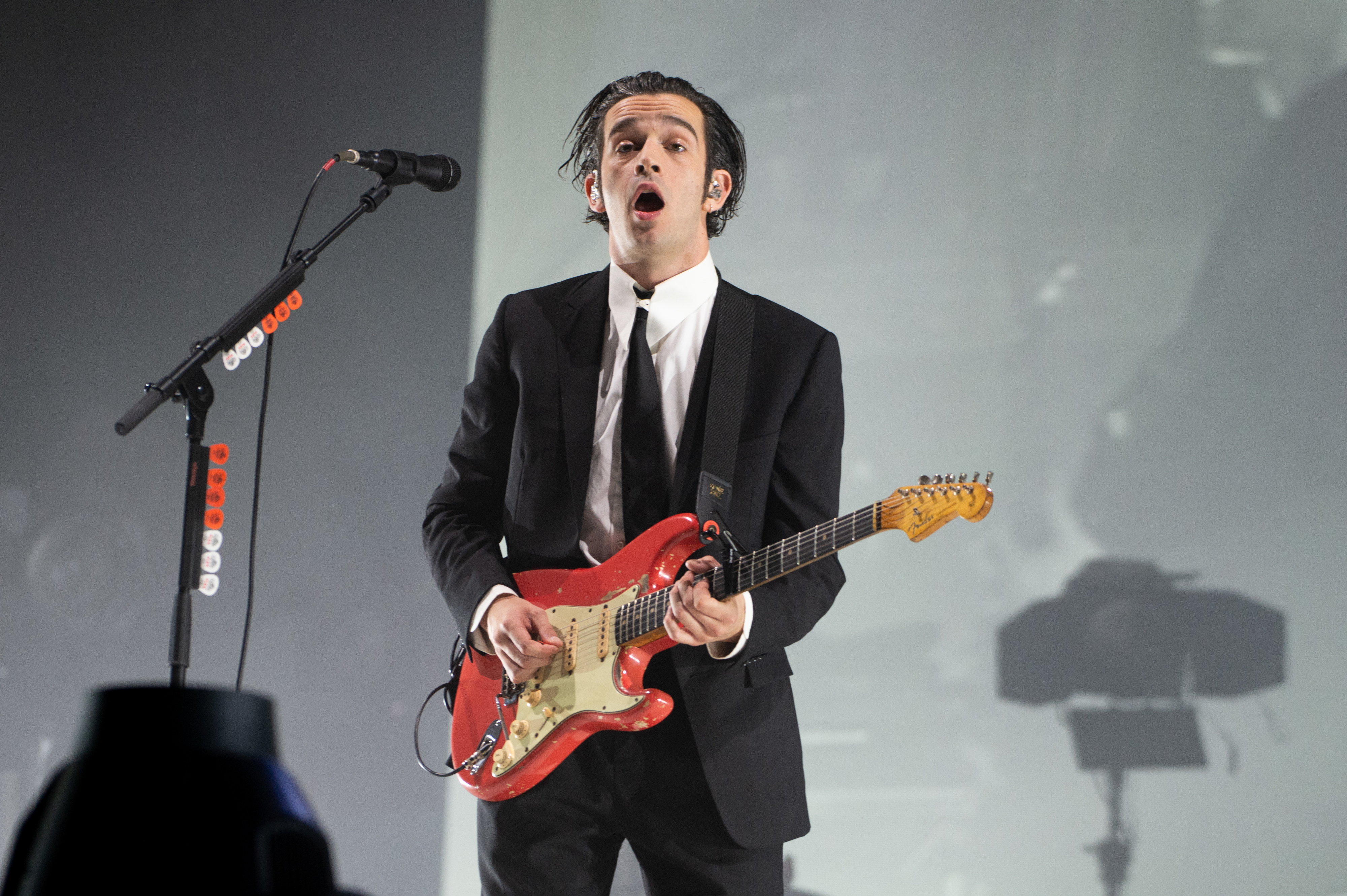 Matty Healy with a guitar wearing a suit and tie performing on stage