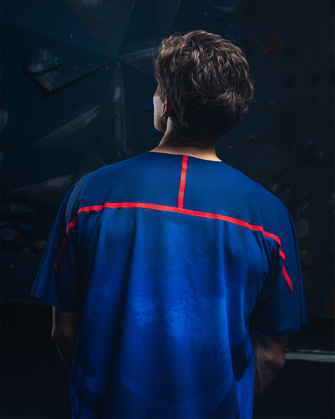 Person in a blue sport shirt with a red stripe from shoulder to shoulder, looking away, in front of a climbing wall