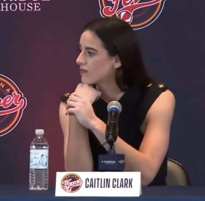 Caitlin Clark seated at a press conference with a microphone in front, wearing a sleeveless top