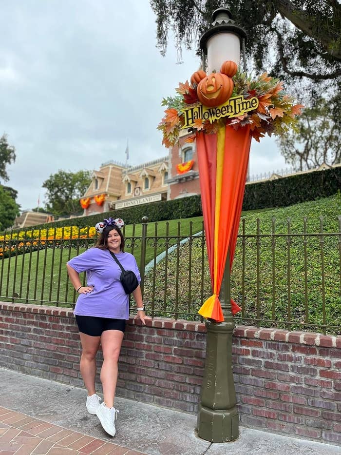 Reviewer posing near a &quot;Halloween Time&quot; sign at Disneyland in the purple tee with shorts and crossbody bag