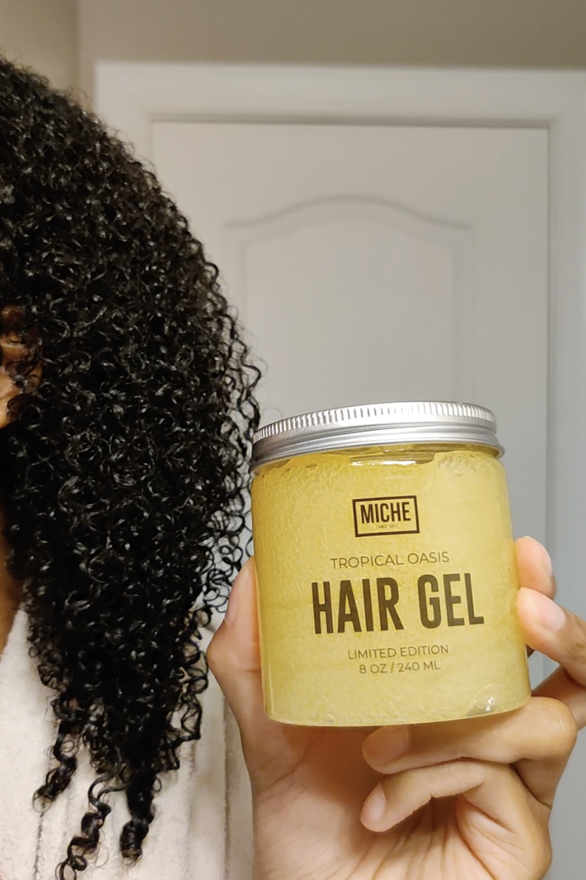 Person holding a jar of Tropical Oasis Hair Gel in front of their curly hair, partly obscuring their face