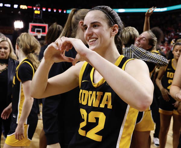Basketball player in Iowa uniform making heart gesture with hands, smiling, others in background
