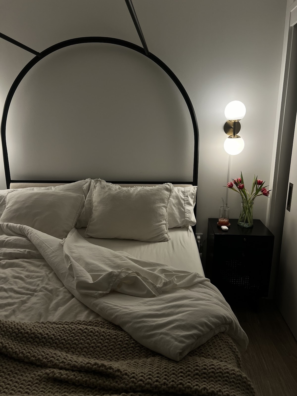 A dimly lit bedroom with an unmade bed, a nightstand with a lamp, and a vase of tulips