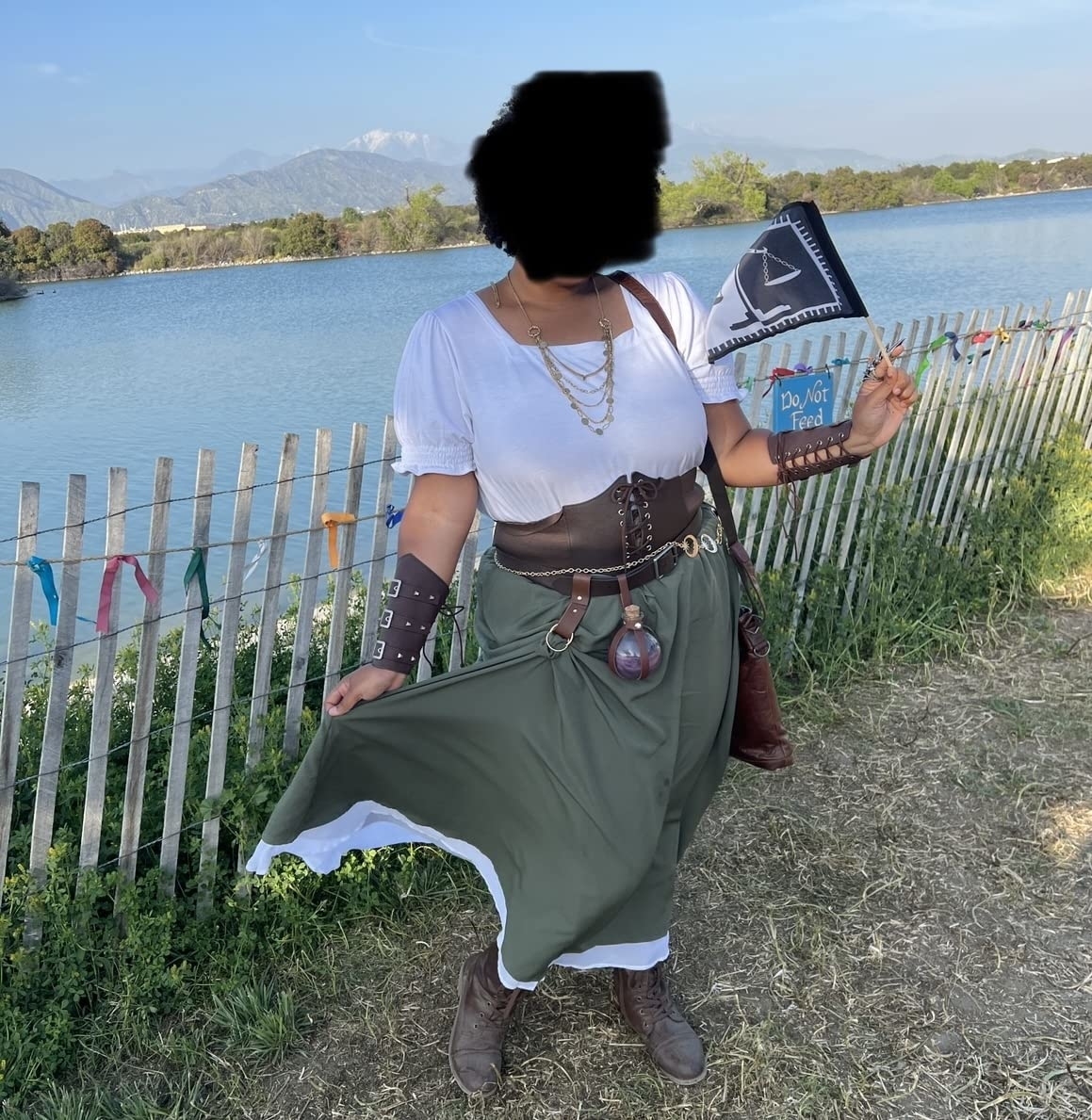 Person in fantasy-inspired outfit with accessories, holding a book, outdoors near a fence
