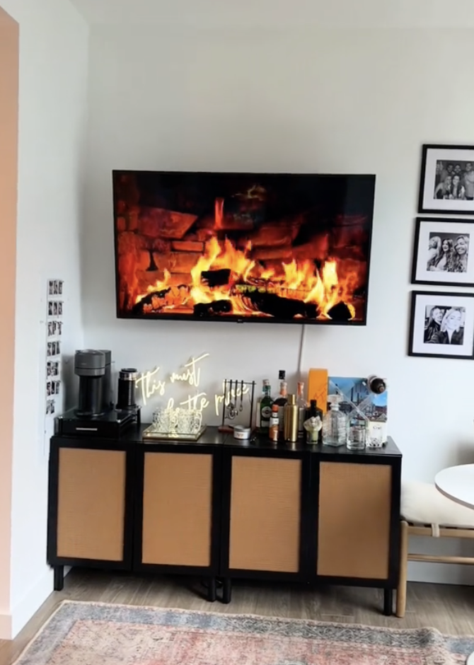 A TV displaying a fireplace video above a cabinet with bottles, a coffee machine, and framed photos on the wall
