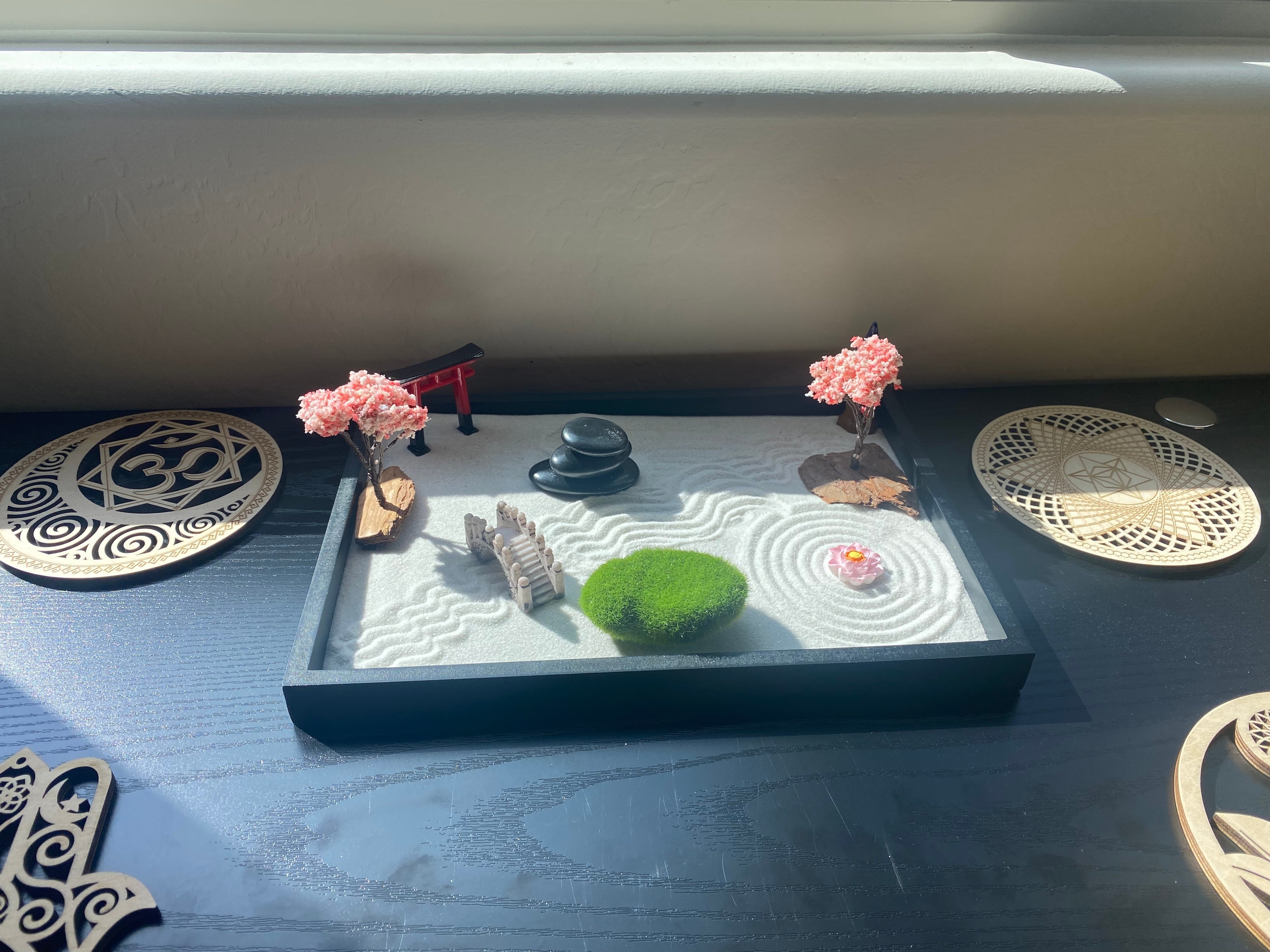 Miniature zen garden with raked sand, decorations, and moss on a sunny windowsill, beside decorative plates. Ideal for mindful home decor