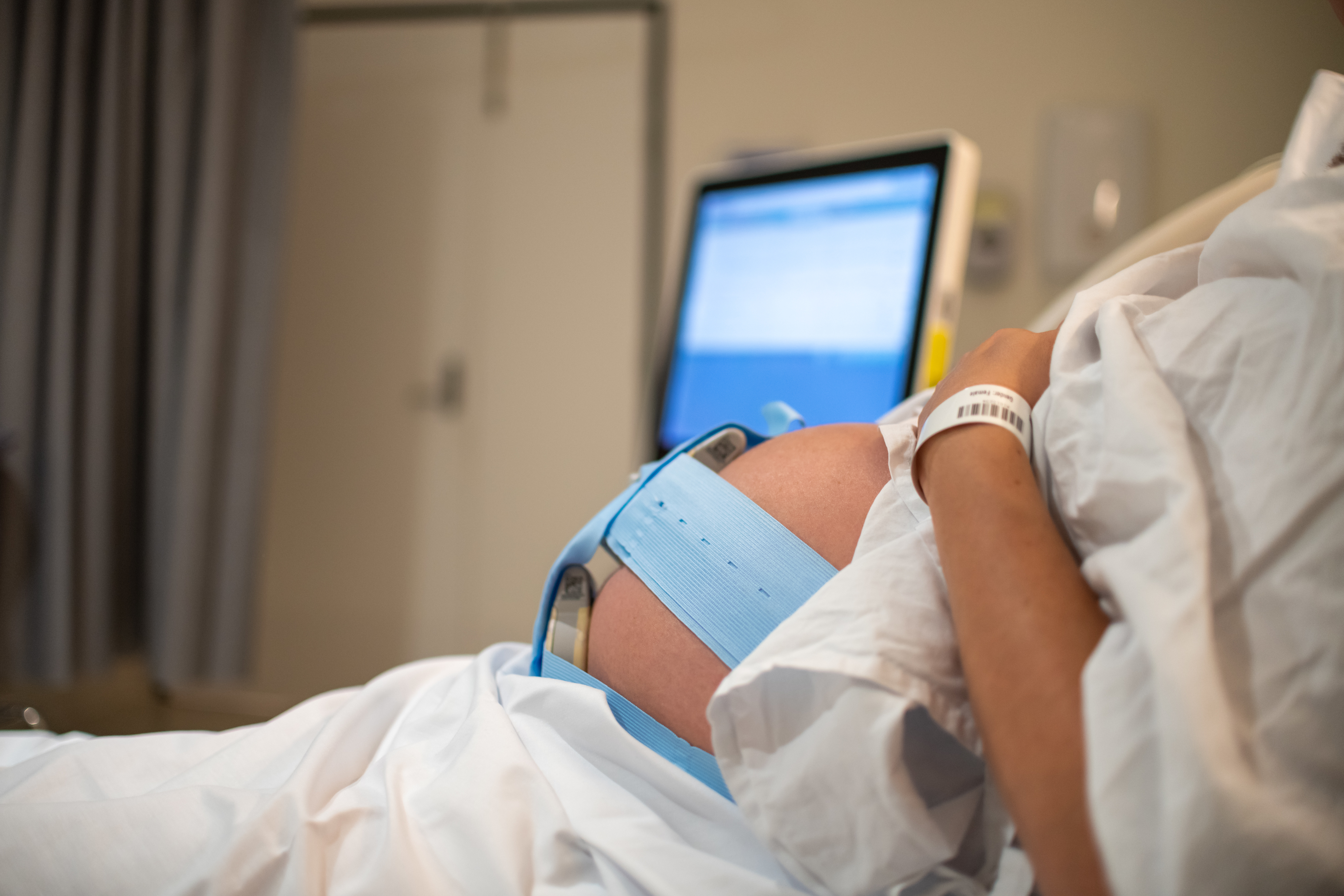 Pregnant person in hospital bed with fetal monitoring bands, monitor in background