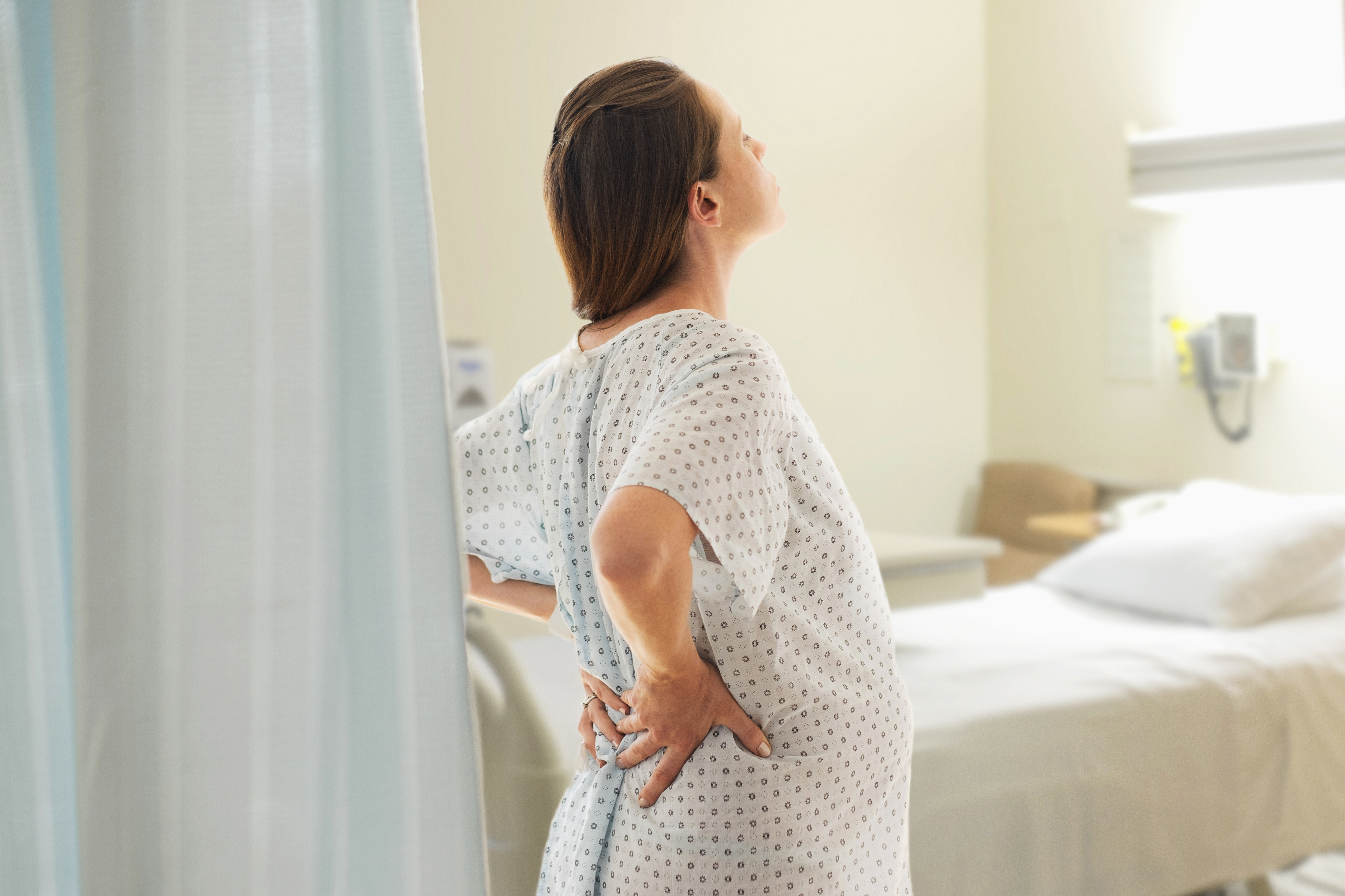 Pregnant person in a hospital gown standing by a curtain, hand on back, expressing discomfort