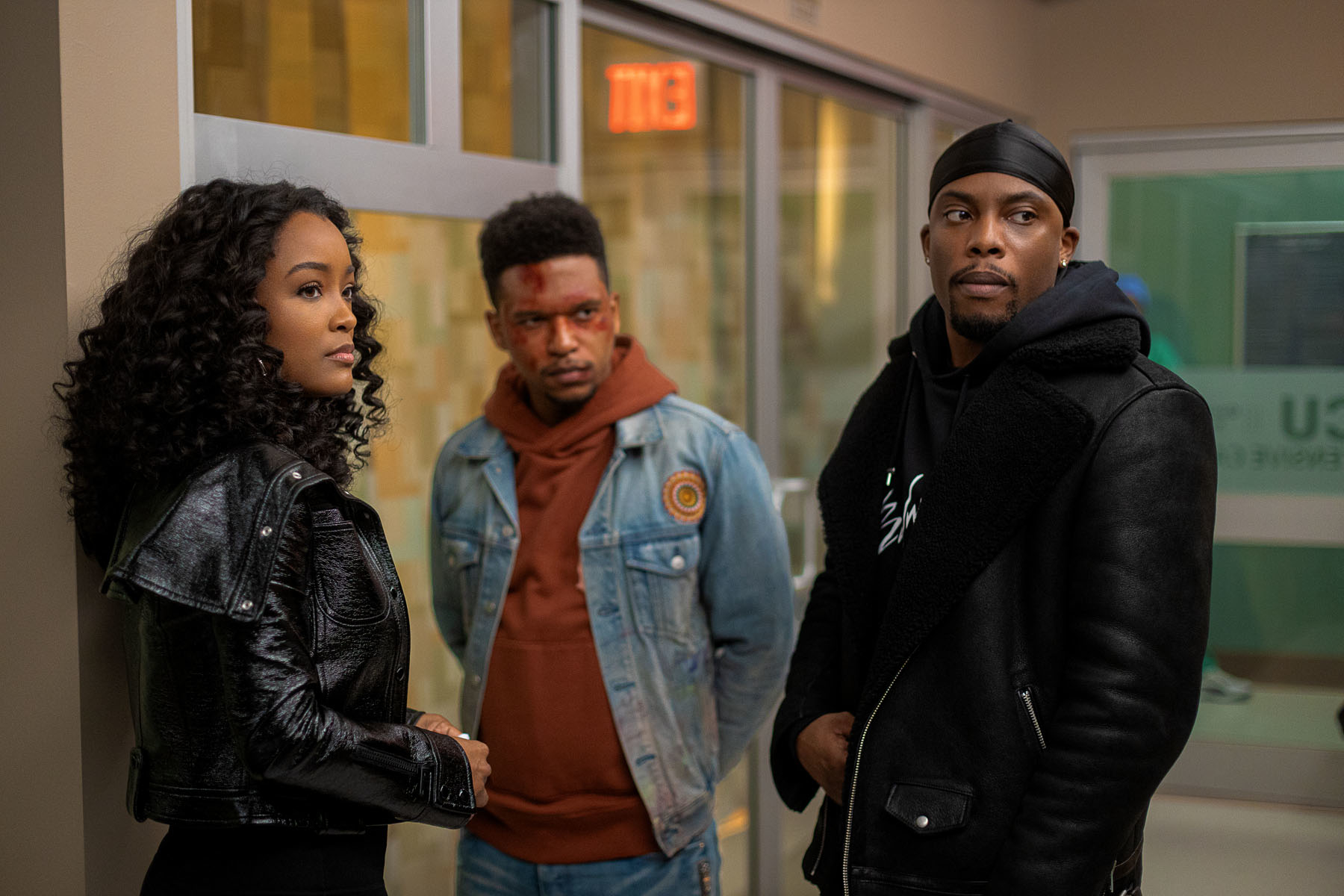 Three actors standing in a hallway, with a man on the right side wearing a hoodie and jacket, and a woman on the left side wearing a leather jacket