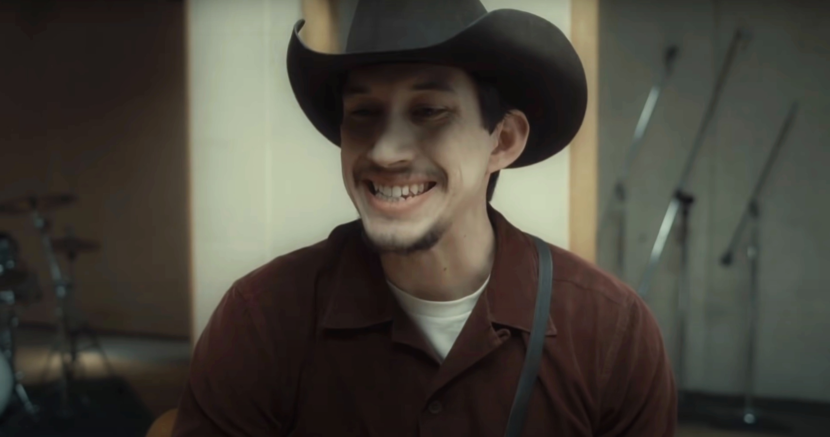 Adam person wearing a cowboy hat and suspenders, sitting indoors and singing