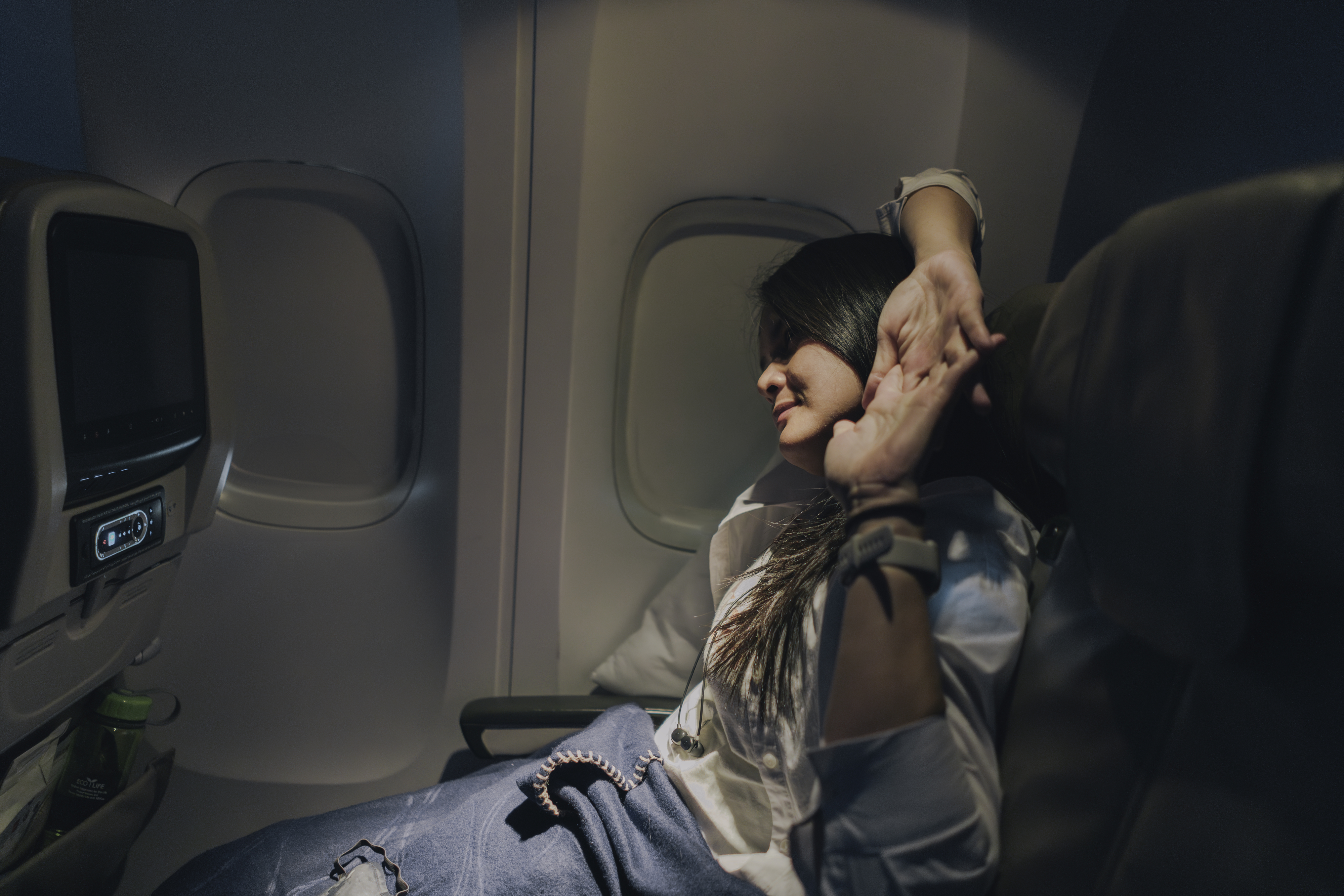 Person relaxing in an airplane seat, stretching arms with closed eyes, in-flight entertainment screen visible