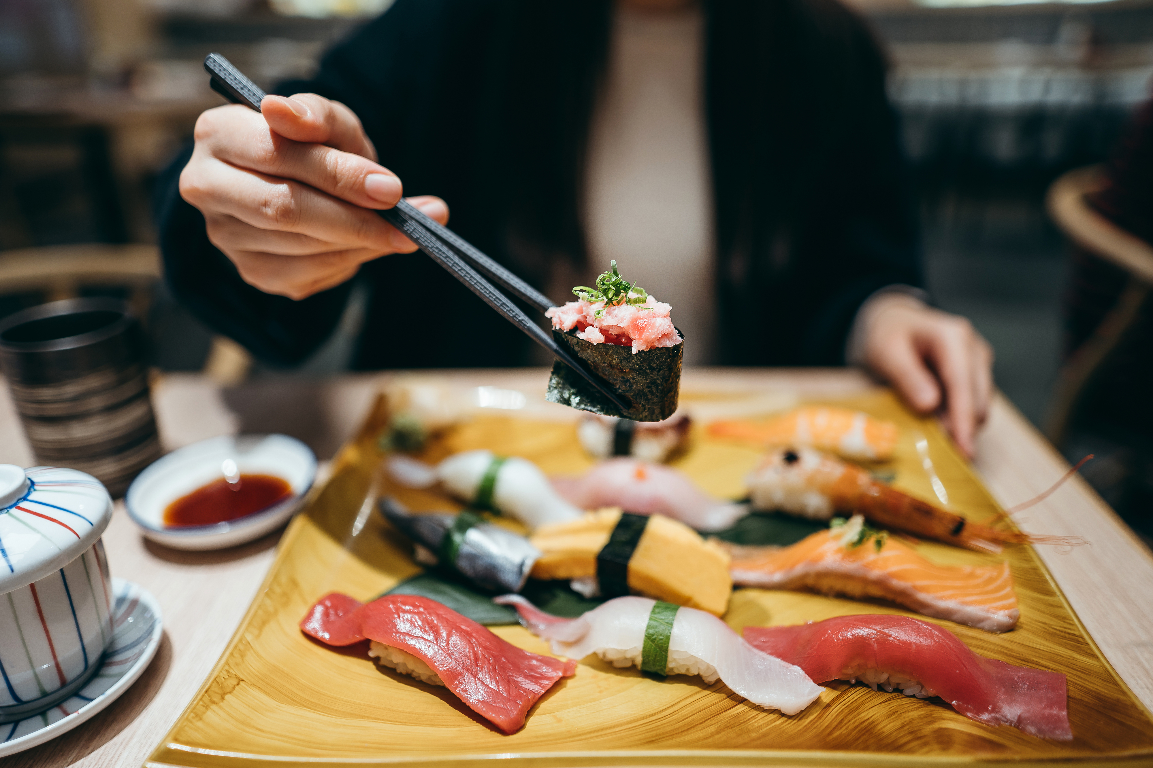 Person holding a piece of sushi with chopsticks over a plate of assorted sushi