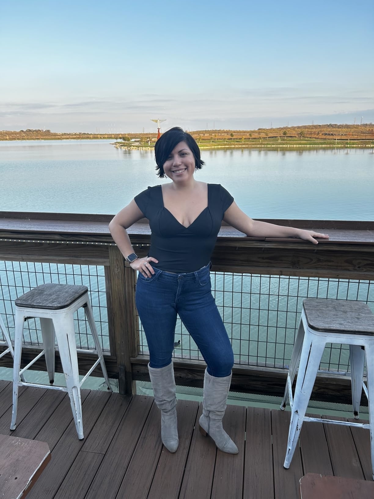 Reviewer in a black corset top, jeans, and knee-high boots poses on a wooden deck with a waterfront view