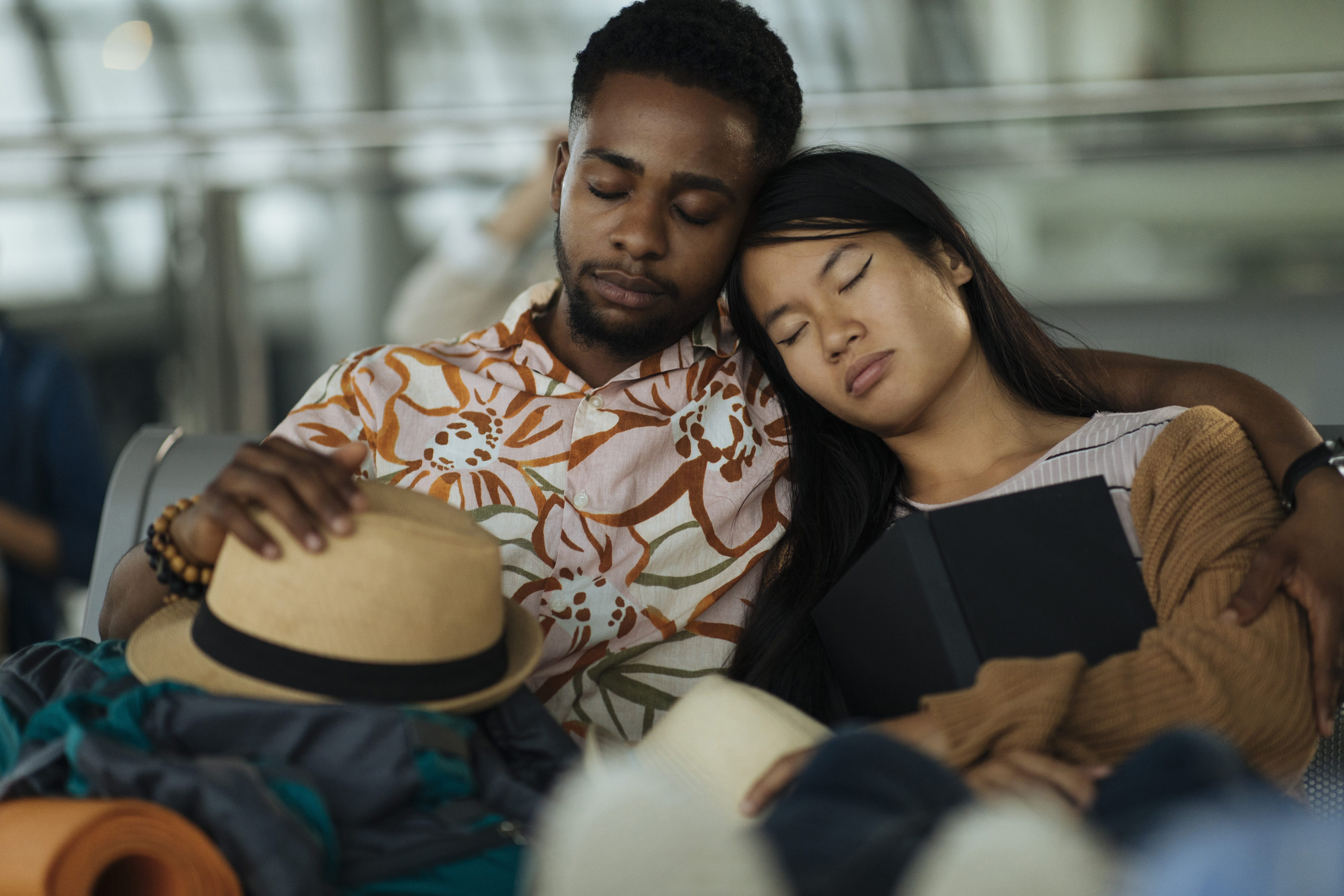 Two travelers rest against each other sleeping in an airport waiting area, surrounded by luggage