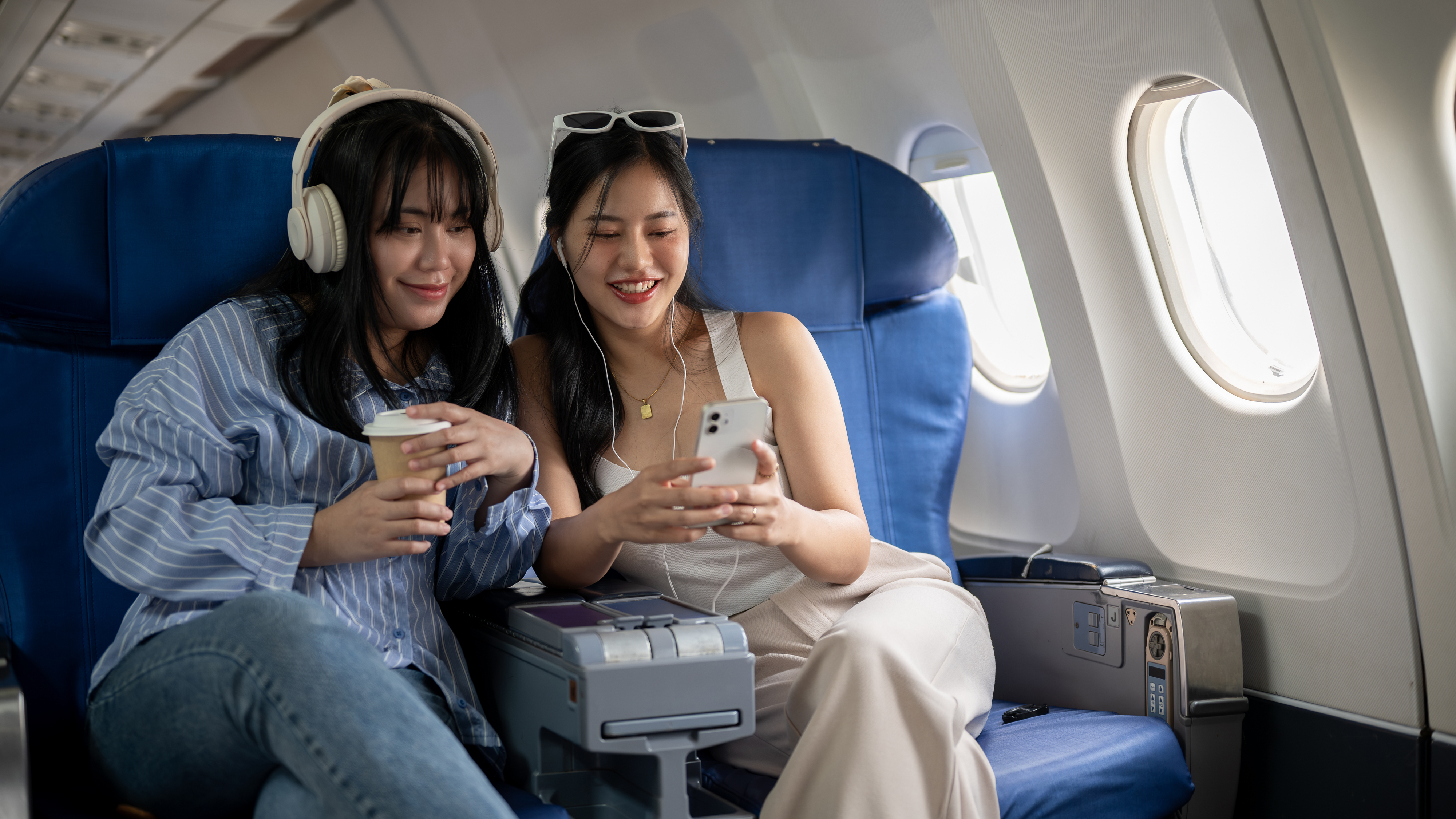 Two women sharing a screen on a phone while seated on a plane, one with headphones
