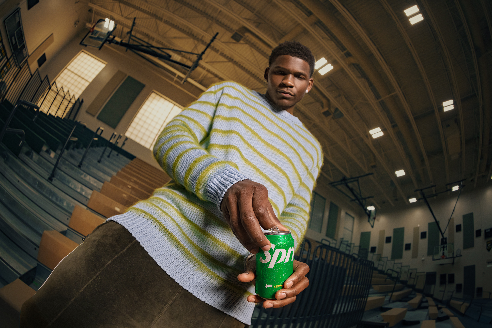 Person sitting in a gym holding a can of Sprite, wearing a striped sweater and leaning on their knee