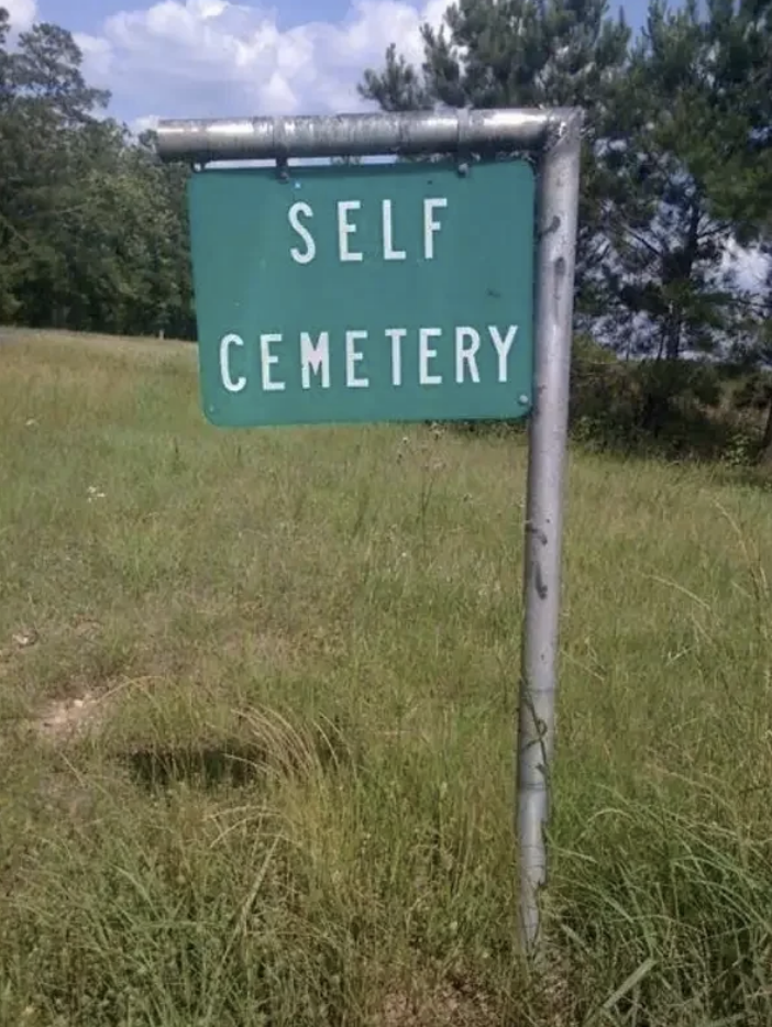 Sign reading &quot;SELF CEMETERY&quot; on a grassy area with trees in the background
