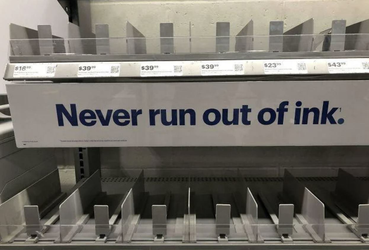Shelves for printer ink cartridges with an empty stock and a sign stating &#x27;Never run out of ink.&#x27;