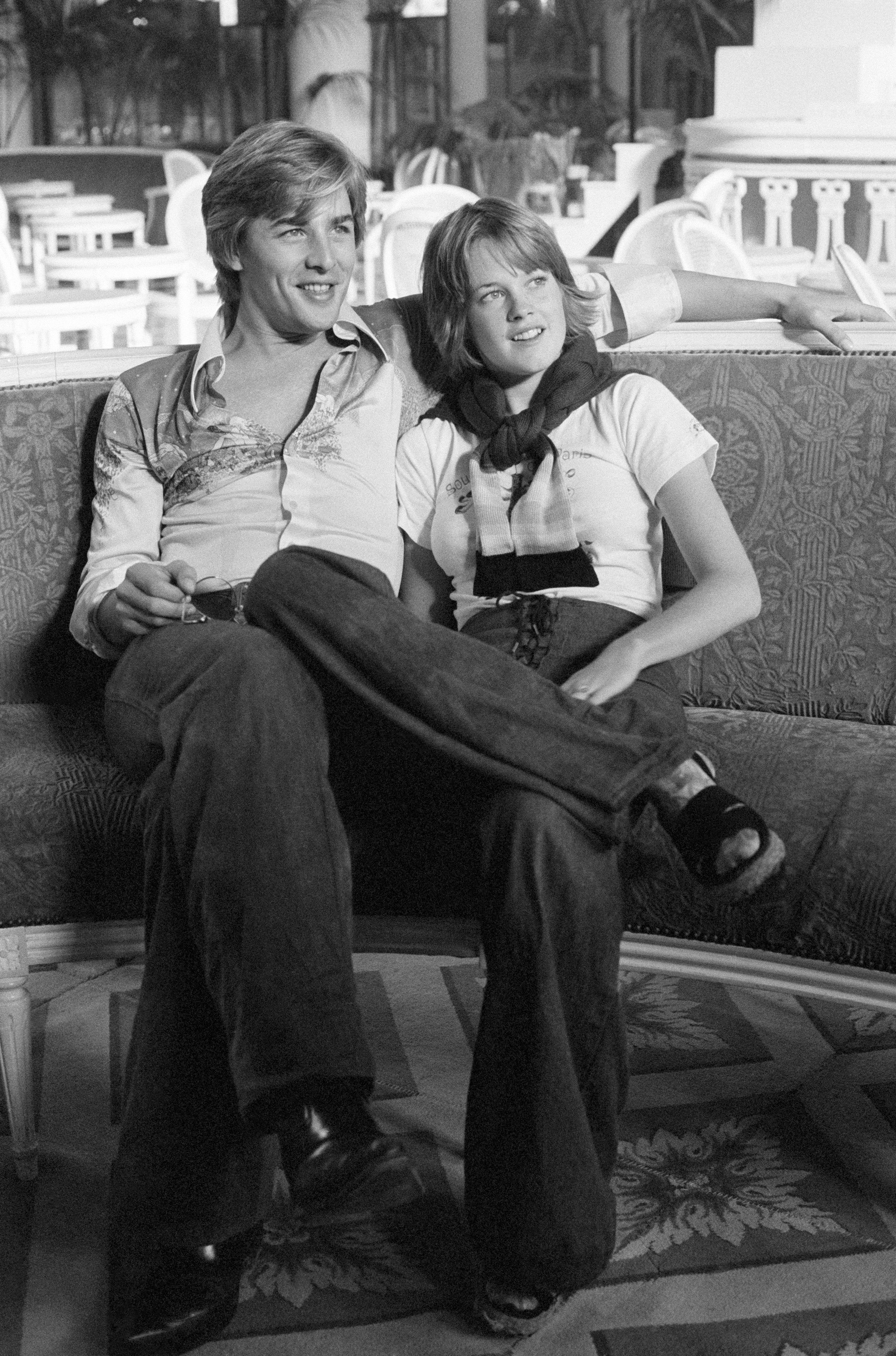 Two people sitting casually, smiling, with one&#x27;s arm around the other. They wear vintage style casual clothing