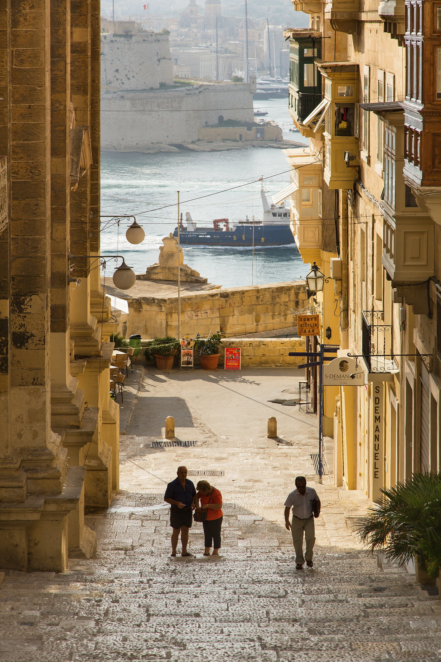 A view down a historic street with three pedestrians and a distant harbor