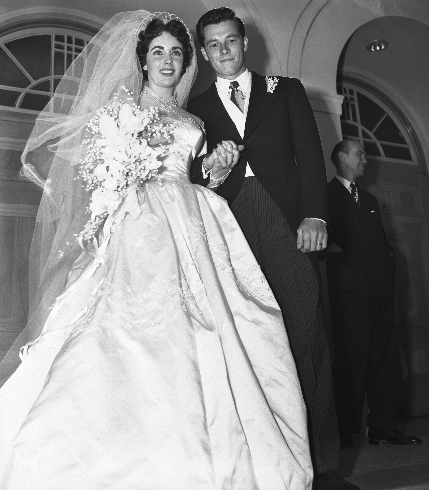 Newlywed couple, bride in a gown with a voluminous veil, groom in a tuxedo, smiling as they walk hand in hand