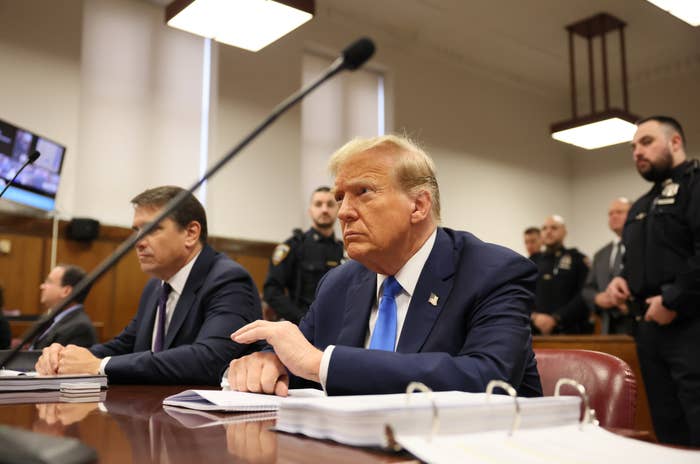 Donald Trump seated at a hearing with serious expression, flanked by officials