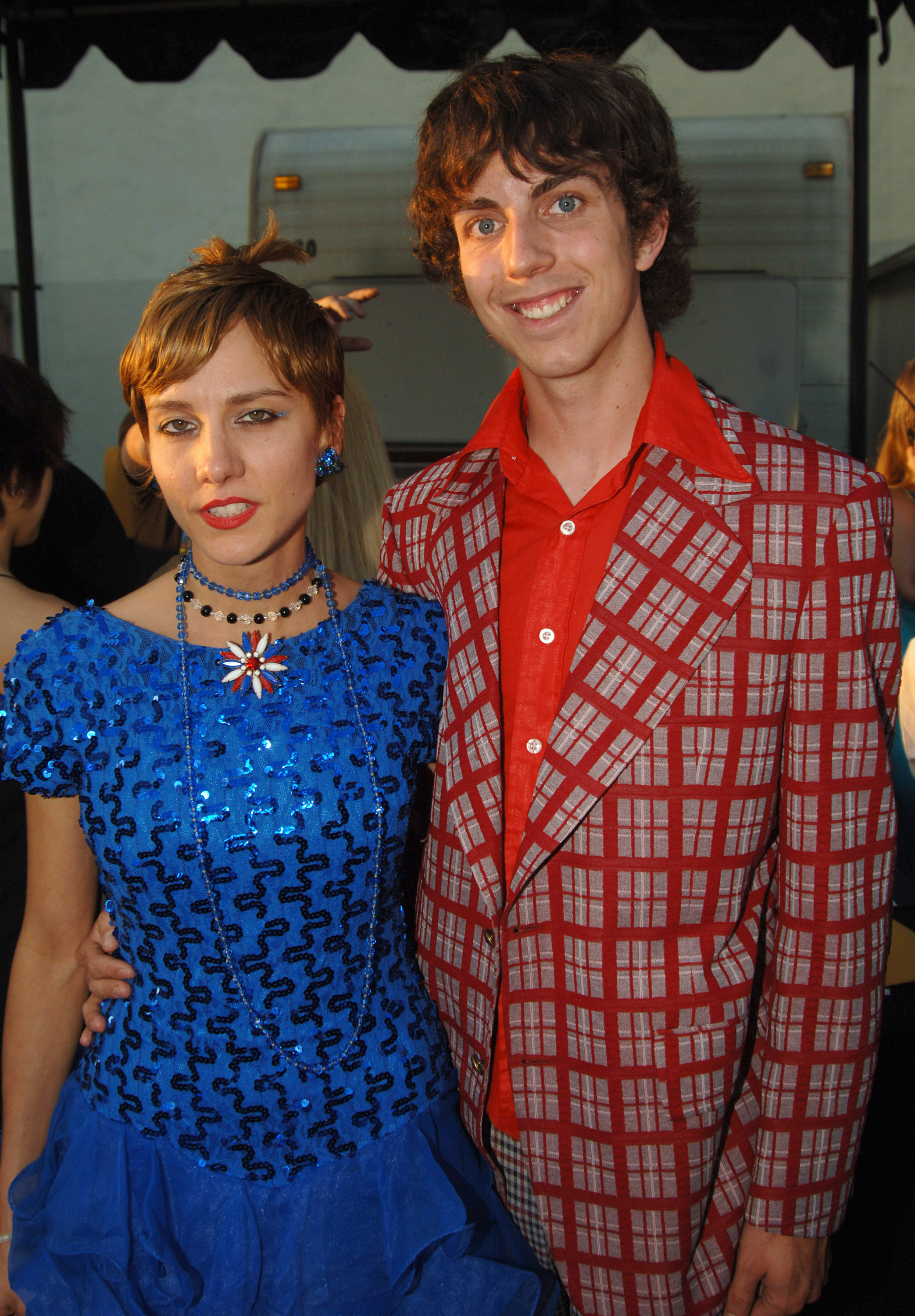 Two people posing; one in a blue sequined dress with an oversized bow, the other in a red checkered suit jacket