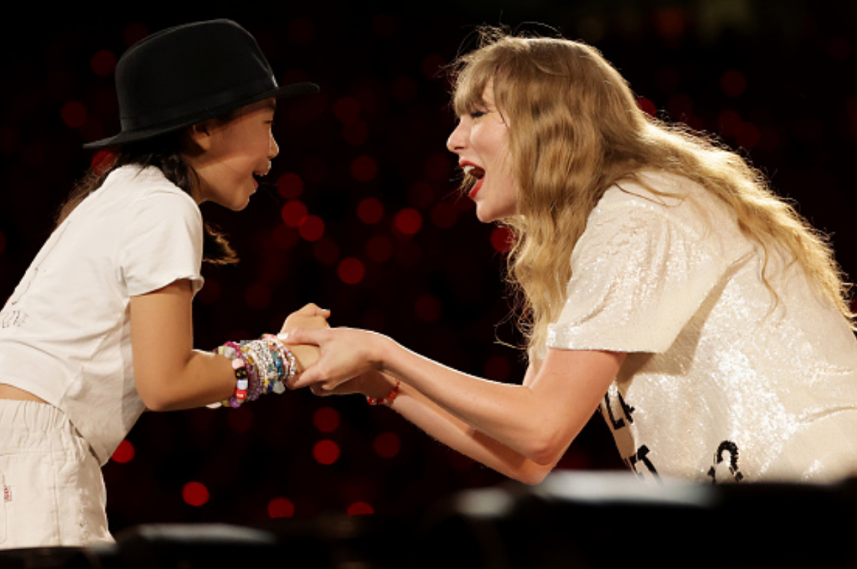 Taylor Swift, in a sequined top, is smiling and holding hands with a young fan wearing a hat onstage
