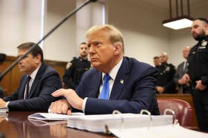 Donald Trump seated at a hearing with serious expression, flanked by officials
