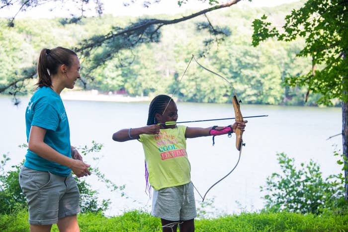 A woman instructs a young girl in archery beside a lake, both in casual attire