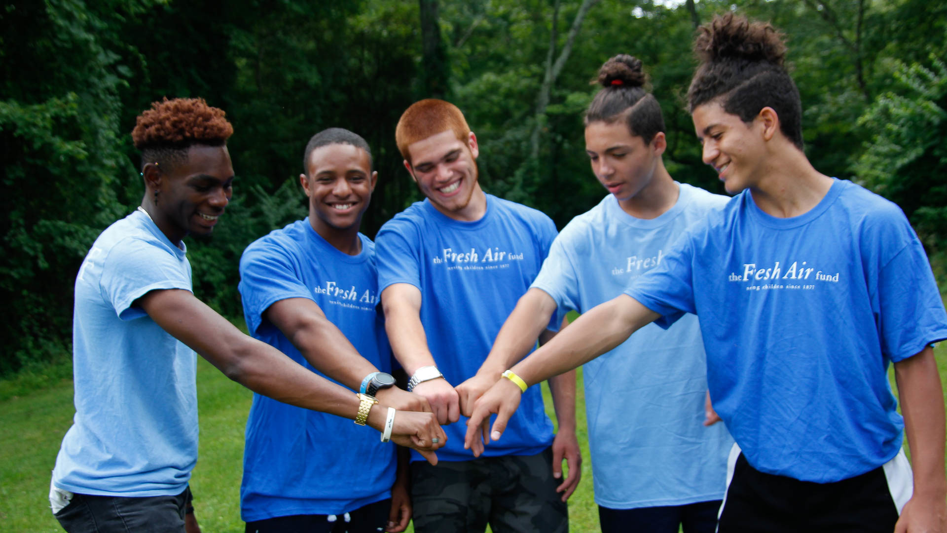 Group of smiling youths in blue Fresh Air Fund t-shirts joining hands in camaraderie