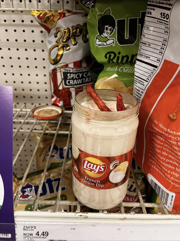Jar of Lay&#x27;s French Onion Dip with two snack sticks in it, on a grocery store shelf with various snack bags around